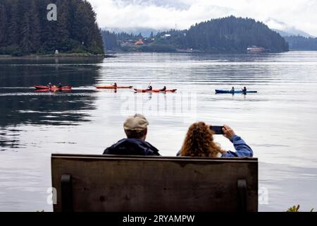 Ein Paar, das auf einer Bank sitzt und Kajakfahrer in Hoonah, Alaska, USA, beobachtet Stockfoto