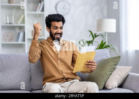 Ein Mann im Haus sitzt auf dem Sofa im Wohnzimmer, erhält einen Briefumschlag, feiert freudig den Sieg. Stockfoto