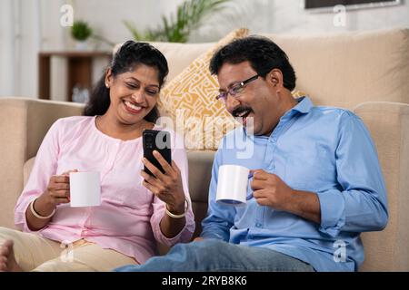 Glückliches, lächelndes indisches Paar mittleren Alters, das Kaffee trinkt, während es zu Hause das Handy benutzt - Konzept der Erfrischung, Social Media Sharing und Stockfoto