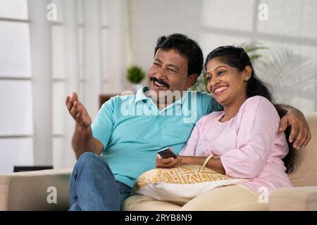 Glücklich lächelndes indisches Paar mittleren Alters, das fernsehen oder Fernsehen schaut, indem es sich unterhält, während es zu Hause auf dem Sofa sitzt - Konzept der Entspannung, Familie Stockfoto