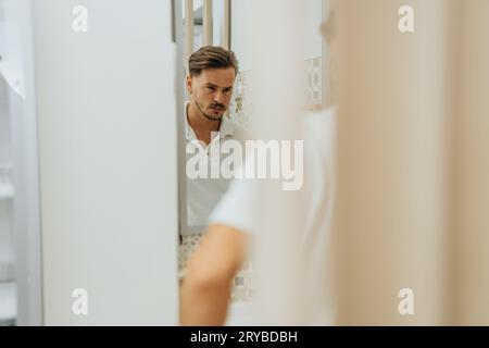 Junger, männlicher, müder und enttäuschter medizinischer Mitarbeiter, der sich in einem Spiegel in der Klinik ansieht. Stockfoto