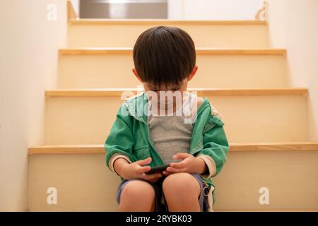 Ein Junge, der von seiner Mutter heimlich beschimpft wurde, kam, um auf der Treppe am Telefon zu spielen. Stockfoto