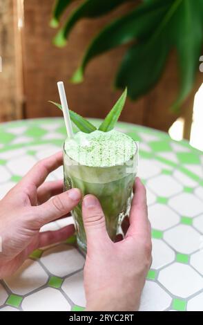 Hände halten Blended Brew Matcha Latte mit Strohhalm auf grünem Tisch und Holzwand Stockfoto