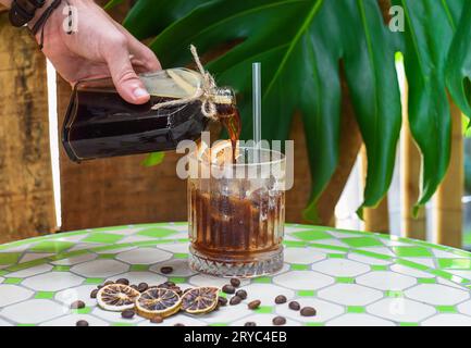 Geben Sie von Hand Kaltkaffee in eine Flasche mit transparentem Glas voller Eis auf grünem Blatthintergrund Stockfoto