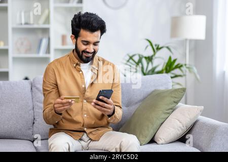 Junger fröhlicher Mann, der zu Hause auf dem Sofa sitzt, die Anwendung am Telefon für Online-Shopping im Online-Shop verwendet, indischer Mann, der zufrieden lächelt und die Kreditkarte hält. Stockfoto