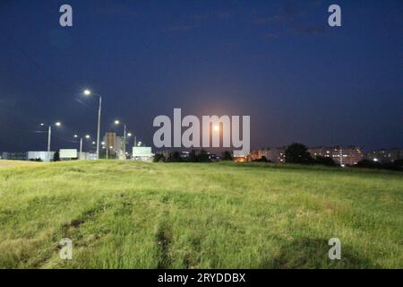 Der Mond am Stadtrand bei Nacht unter dem Licht der Laternen. Nächtliche Stadtlandschaft Mond über Gras. Stockfoto