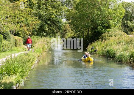 Ein Mann, der im August einen Kanalweg entlang geht, mit einem Mann und einer Frau, die im Kajak auf dem Basingstoke-Kanal bei Odiham paddeln. Hampshire, England Stockfoto