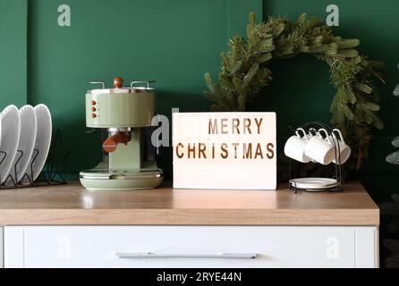 Brett mit Text FROHE WEIHNACHTEN, Kaffeemaschine und Kranz auf Küchentheke in der Nähe der grünen Wand Stockfoto
