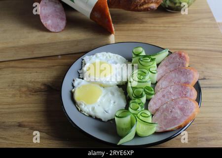 Frühstück traditionelles Rührei Spiegeleier zwei Schinkenwürste und Gurkensalat auf einem grauen Teller auf dem Tisch. Daneben befinden sich Produkte zum Kochen Stockfoto