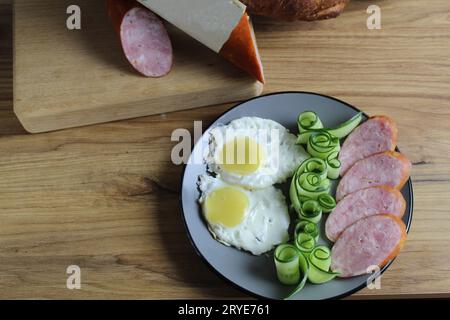 Frühstück traditionelles Rührei Spiegeleier zwei Schinkenwürste und Gurkensalat auf einem grauen Teller auf dem Tisch. Daneben befinden sich Produkte zum Kochen Stockfoto