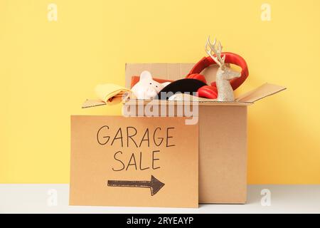 Schachtel mit unerwünschten Sachen und Pappschild mit Text GARAGE SALE auf GELBEM Hintergrund Stockfoto