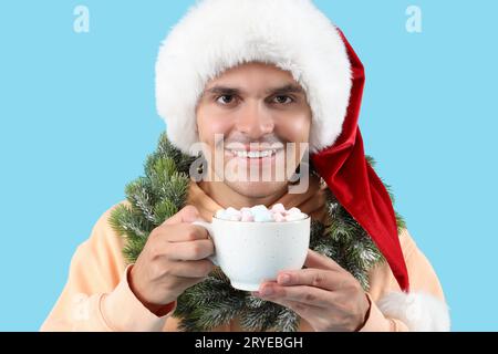 Glücklicher junger Mann im Weihnachtsmannhut mit Weihnachtskranz und Tasse Kakao auf blauem Hintergrund Stockfoto