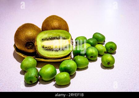 Drei Kiwi-Früchte, eines in zwei Hälften auf goldenem Metallblatt geschnitten, umgeben von einem Haufen Mini-Kiwi isoliert Stockfoto