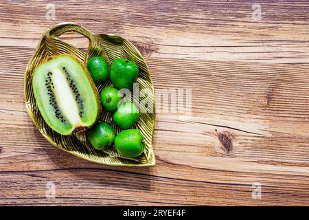 Ein Haufen Mini-Kiwis mit einer normalen Kiwi, die auf einer goldenen Blattplatte aus Metall in zwei Hälften geschnitten wurde, liegen flach auf einem hölzernen Hintergrund Stockfoto