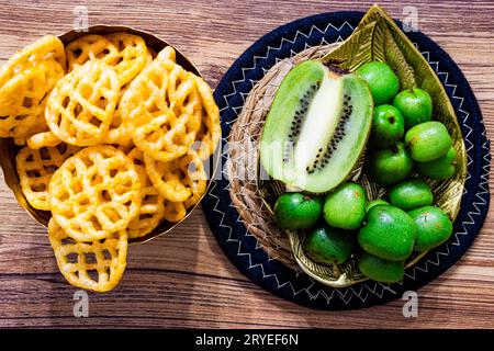 Ein Haufen Mini-Kiwis mit einer normalen Kiwi, die auf einer goldenen Blattplatte aus Metall auf einer runden Strohmatte mit einer Schüssel gebratener Chips Health in der Mitte geschnitten wird Stockfoto