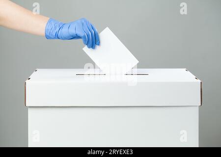 Kaukasische Frauenhand mit blauem Latex-Gummihandschuh, der Stimmzettel in die Wahlbox legt Stockfoto