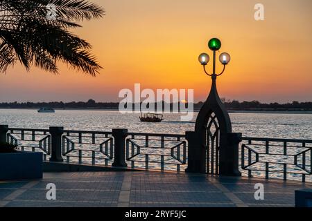 Sonnenuntergang in Abu Dhabi Corniche mit einem traditionellen, silhouettierten Boot, das vorbeifährt Stockfoto