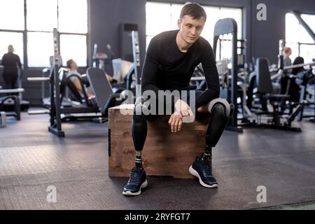 Porträt in voller Länge eines ernsthaften Sportlers mit Beinprothese, der auf der Fitnessbox sitzt und nach dem Workout, Freizeit, Freizeit. Lebensweise, Müdigkeit Stockfoto