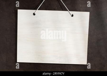 Weißes, weißes Holzschild, das an einer dunklen strukturierten Wand hängt. Mustervorlage Stockfoto