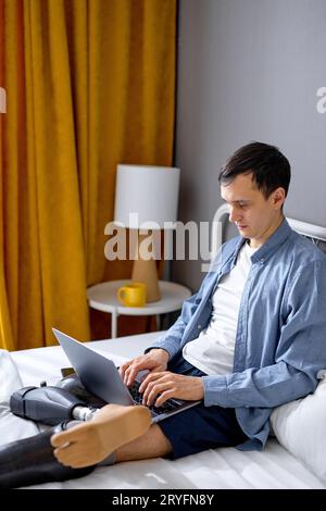 Konzentrierter junger Mann mit Prothese auf Beinen mit Laptop für Fernarbeit zu Hause, sitzend auf dem Bett. Selbstbewusster Kaukasier mit körperlicher Behinderung h Stockfoto