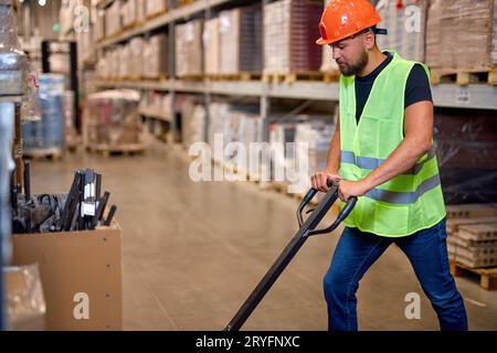 Männliche Mitarbeiter im Lager verwenden Handhubwagen, um Waren zu transportieren, allein, in Arbeitskleidung und Schutzhelm gekleidet. Fachlager EM Stockfoto