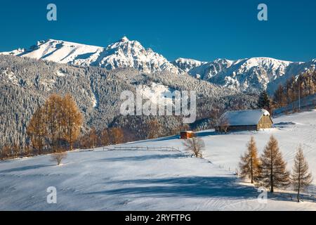 Malerische Winterlandschaft mit schneebedeckter Holzhütte am Hang und spektakulären hohen Bergen im Hintergrund, Bucegi-Bergen, Karpaten, Roma Stockfoto