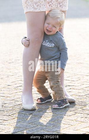 Trauriger kaukasischer blonder kleiner Junge, der ihre Mutter an den Beinen umarmt Stockfoto