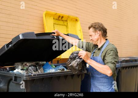 Kaukasischer gutaussehender Mann Angestellter öffnet Dose voller Müll, um Plastiktüte mit Schürze zu werfen. Falscher unsortierter Müll c Stockfoto