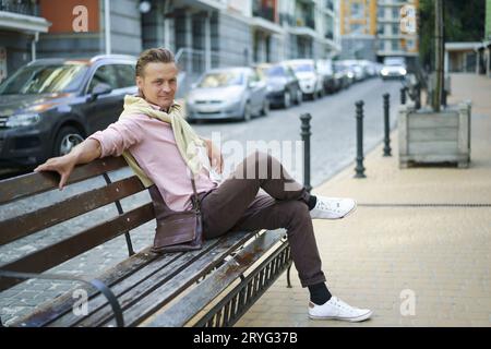 Ein gutaussehender Mann sitzt mit überkreuzten Beinen auf der Bank und verbringt Zeit in der Stadt in der Nähe seines Büros oder Hauses, trägt ein rosafarbenes Hemd und sollte es sein Stockfoto