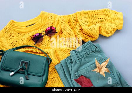 Flaches, gelbes Damenpullover, grüne Jeans, Tasche und Sonnenbrille, Herbstblätter auf grauem Hintergrund. Modekonzept für den Herbst. Draufsicht, flach. Stockfoto