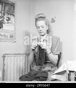 In den 1940er Jahren Eine junge Frau sitzt in der Küche auf einem Stuhl und strickt einen Pullover. In ihrem Schoß sieht man fertige Strickteile des Pullovers und eine Garnkugel. Vielleicht strickt sie ein Kleidungsstück, um es als Weihnachtsgeschenk zu verschenken, wenn eine Weihnachtsdekoration an der Wand neben ihr hängt, die frohe Weihnachten wünscht. und dann muss sie es eilig gehabt haben, rechtzeitig fertig zu werden, um den Pullover als Weihnachtsgeschenk zu verschenken. Schweden 1947. Kristoffersson ref AB22-3 Stockfoto