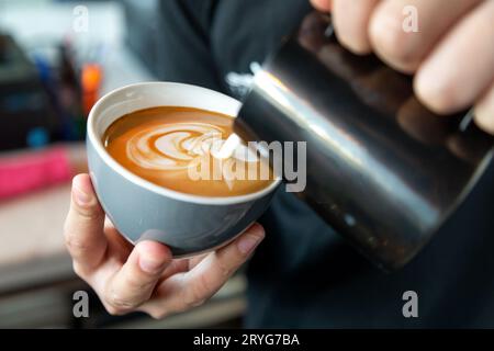 Der Barista bereitet Café Latte zu und gießt Milch in einen Kaffee. Nahaufnahme auf männlichen Händen mit Latte Art. Stockfoto