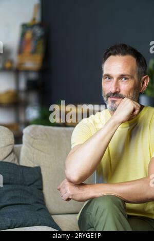 Porträt eines gutaussehenden freiberuflichen Mannes mittleren Alters, der auf dem Sofa sitzt, mit einem Kinn, der seine Hand beugt und in die Kamera blickt, in Casua Stockfoto