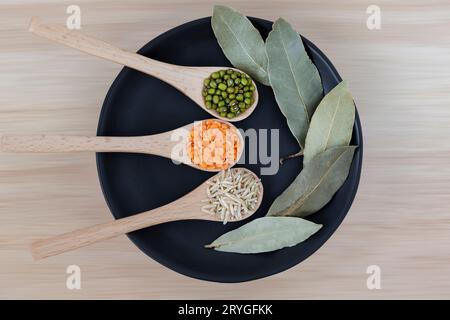 Reis, Mongbohnen und Dal in Holzlöffeln neben Sternaniskernen, Lorbeerblättern und bunten Gewürzen Stockfoto