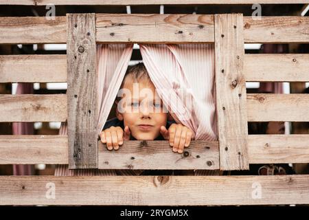 Der süße kleine Junge schaut aus dem Fenster des hölzernen Kinderhauses. Ferien im Dorf. Glückliche Kindheit. Spiele und Unterhaltung für Kinder. Kinderemot Stockfoto