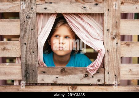 Das süße kleine Mädchen schaut aus dem Fenster des hölzernen Kinderhauses. Ferien im Dorf. Glückliche Kindheit. Spiele und Unterhaltung für Kinder. Kinderemo Stockfoto