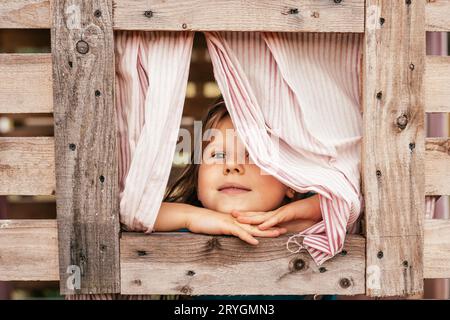 Glückliches kleines Mädchen schaut aus dem Fenster des hölzernen Kinderhauses. Ferien im Dorf. Glückliche Kindheit. Spiele und Unterhaltung für Kinder. Kinder-EM Stockfoto