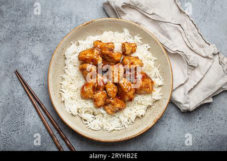 Chinesisches süß-saures, klebriges Huhn mit Sesamsamen und Reis auf Keramikplatte mit Essstäbchen von oben, graues rustikales Steinbaa Stockfoto