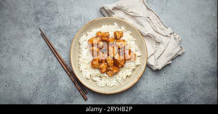 Chinesisches süß-saures, klebriges Huhn mit Sesamsamen und Reis auf Keramikplatte mit Essstäbchen von oben, graues rustikales Steinbaa Stockfoto