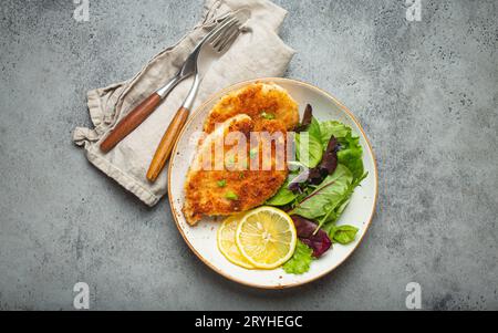 Knuspriges panko paniertes Hähnchenfilet mit grünem Salat und Zitrone auf Teller mit Besteck auf grauem rustikalem Betonhintergrund ta Stockfoto