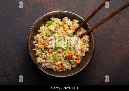Authentischer chinesischer und asiatischer gebratener Reis mit Ei und Gemüse in brauner Keramikschale mit Blick von oben, dunkler rustikaler Betontisch Backgr Stockfoto