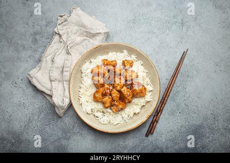 Chinesisches süß-saures, klebriges Huhn mit Sesamsamen und Reis auf Keramikplatte mit Essstäbchen von oben, graues rustikales Steinbaa Stockfoto