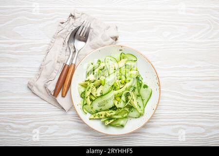 Gesunde vegane grüne Avocado Salatschüssel mit geschnittenen Gurken, Edamame Bohnen, Olivenöl und Kräutern auf Keramikplatte Draufsicht, weiß Stockfoto