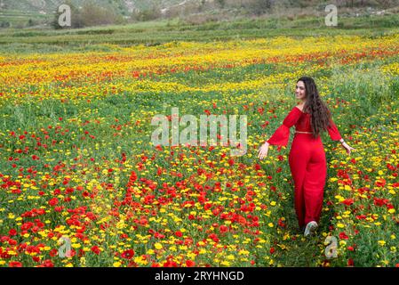 Attraktive junge Frau in rotem Kleid, die glücklich im Frühlingsfeld mit Mohnblumen läuft. Stockfoto