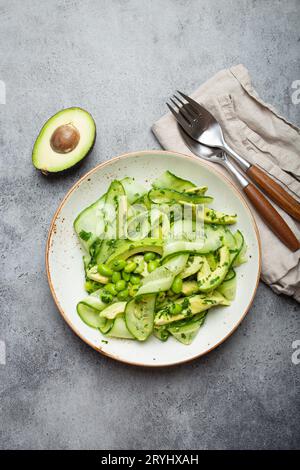 Gesunde vegane grüne Avocado Salatschüssel mit geschnittenen Gurken, Edamame Bohnen, Olivenöl und Kräutern auf Keramikplatte Draufsicht, grau Stockfoto