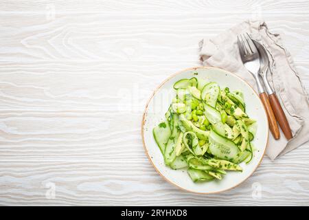 Gesunde vegane grüne Avocado Salatschüssel mit geschnittenen Gurken, Edamame Bohnen, Olivenöl und Kräutern auf Keramikplatte Draufsicht, weiß Stockfoto