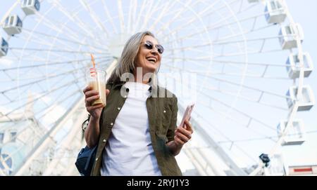 Fröhliche Seniorin genießt einen sonnigen Tag in einer alten europäischen Stadt, hält ihr Telefon und eine Tasse Saft in ihren Händen mit einem großen s Stockfoto