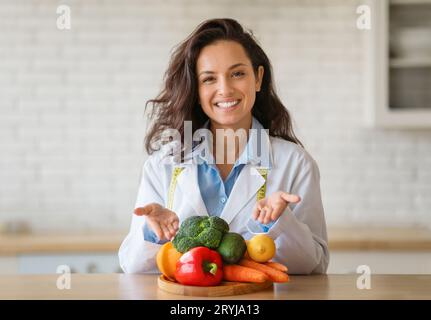 Porträt eines fröhlichen Diätassistenten im Labormantel, der auf frisches Obst und Gemüse auf dem Tisch zeigt und in der Klinik vor der Kamera lächelt Stockfoto