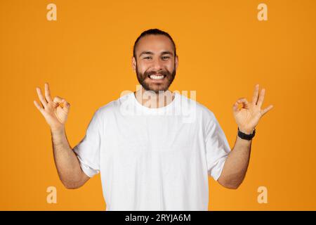 Lächelnder erwachsener europäischer Mann mit Bart in weißem T-Shirt, der mit den Händen ein gutes Zeichen macht Stockfoto