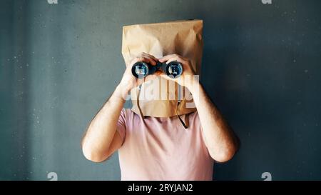 Mann mit einer Papiertüte auf dem Kopf, der durch das Fernglas zur Kamera schaut Stockfoto